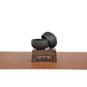 Two handcrafted Jianshui zitao tea cups with a matte black finish, displayed on a carved wooden stand