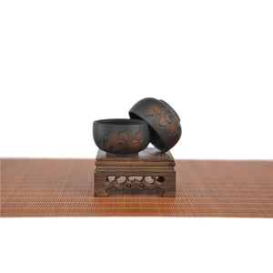 Two Jianshui zitao black cups with line-carved floral patterns, displayed on a carved wooden stand