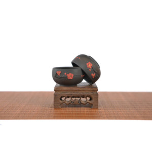 Two handcrafted Jianshui zitao tea cups with plum blossom motif, displayed on a carved wooden stand