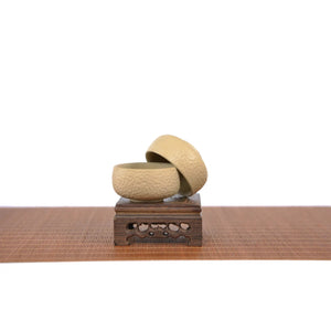Two handcrafted white clay teacups with a hammered texture, displayed stacked on a carved wooden stand, highlighting the unglazed matte surface and artisanal finish.