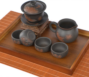 Hand-carved lotus tea set arranged on a wooden tea tray, including a lotus-engraved gaiwan on a stand, a matching fairness pitcher, and four dark clay teacups with carved lotus motifs.