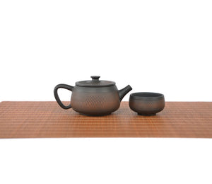 Handcrafted pottery teapot with a chattered knife texture and unglazed matte finish, paired with a matching teacup on a woven tea mat.