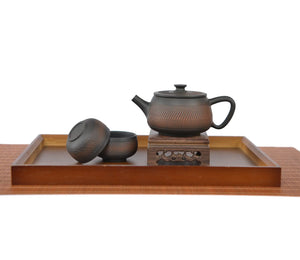 Handcrafted pottery teapot with a chattered knife (jump-cut) texture and unglazed matte finish, displayed on a carved wooden stand with two matching teacups on a tea tray.