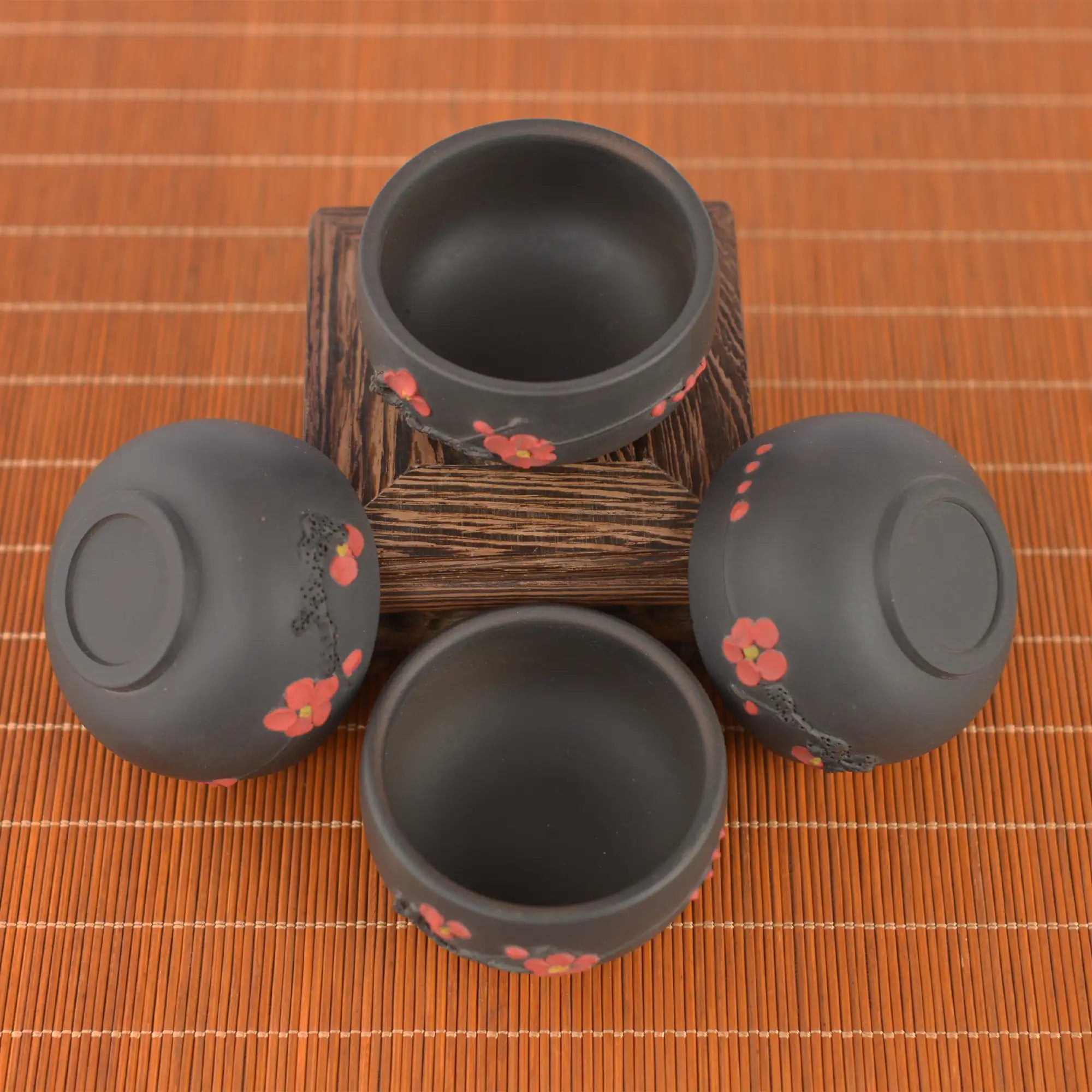 Four Jianshui zitao tea cups with red plum blossom accents, arranged on a wooden stand over a bamboo mat