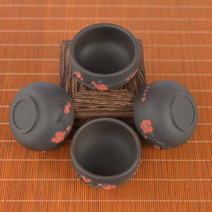 Four Jianshui zitao tea cups with red plum blossom accents, arranged on a wooden stand over a bamboo mat