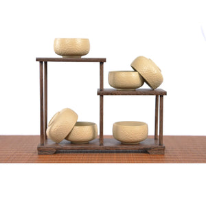 Six handcrafted white clay teacups with a hammered texture displayed on a two-tier wooden stand, showing multiple angles and the unglazed matte finish of the minimalist tea cup set.