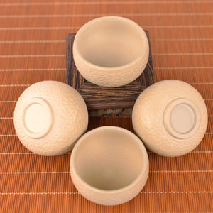 Four handcrafted white clay teacups with a hammered texture, shown from a top view on a bamboo mat and wooden stand, highlighting the unglazed matte finish and minimalist design.