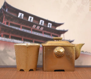 Handcrafted wood-fired clay square teapot and matching cup set, displayed together on a woven mat