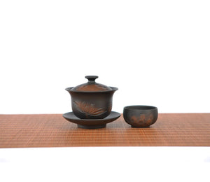 Hand-carved lotus gaiwan set with one matching teacup, featuring fine line-engraved lotus motifs on dark clay, displayed on a bamboo mat.