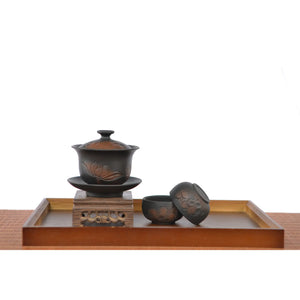 Hand-carved lotus gaiwan set displayed on a wooden tray, featuring a lotus-engraved gaiwan on a stand with two matching dark clay teacups placed beside it.