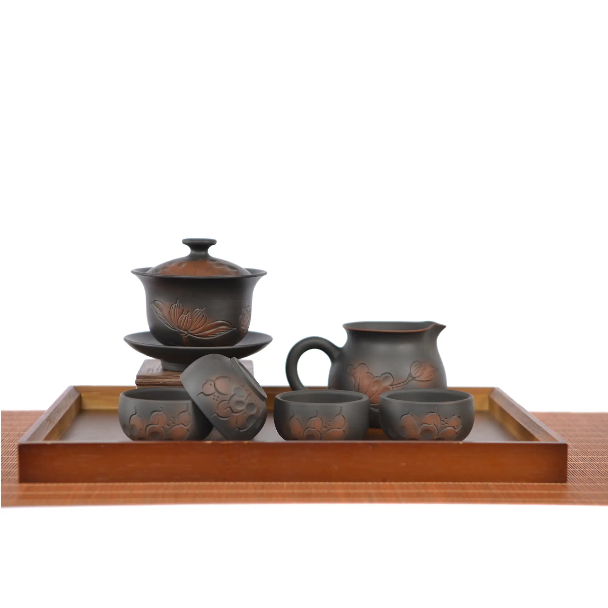 Hand-carved lotus tea set displayed on a wooden tray, featuring a lotus-engraved gaiwan on a stand, a matching fairness pitcher, and four dark clay teacups arranged neatly in front.