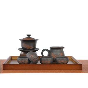 Hand-carved lotus tea set displayed on a wooden tray, featuring a lotus-engraved gaiwan on a stand, a matching fairness pitcher, and four dark clay teacups arranged neatly in front.