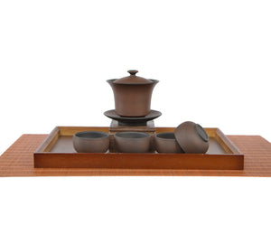 Warm brown Bodhi-pattern pottery gaiwan with saucer displayed on a carved wooden stand, paired with four matching teacups arranged on a rectangular tea tray, with one cup turned on its side